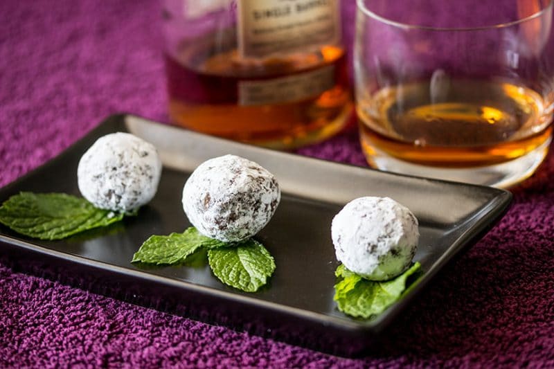 Powder sugar coated bourbon balls and mint leaves on a plate plate with bourbon in the background