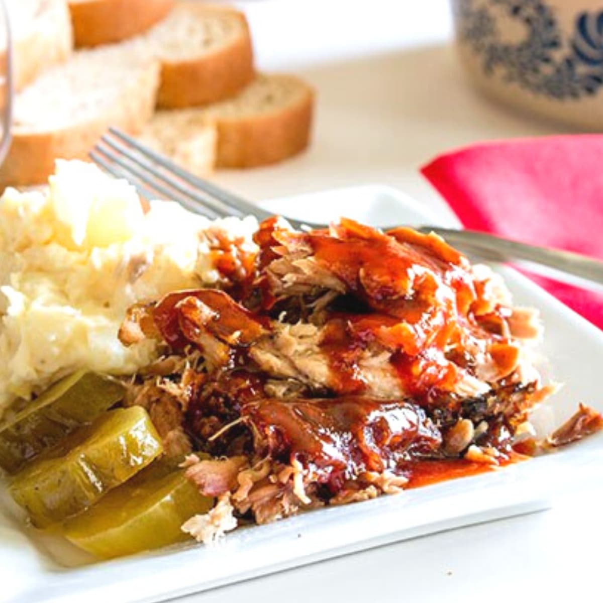 Pulled pork covered in BBQ auce on a plate with pickles and potato salad