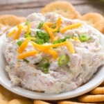 Ham and cream cheese dip topped with cheddar cheese shreds and green onion slices in a white bowl surrounded by crackers