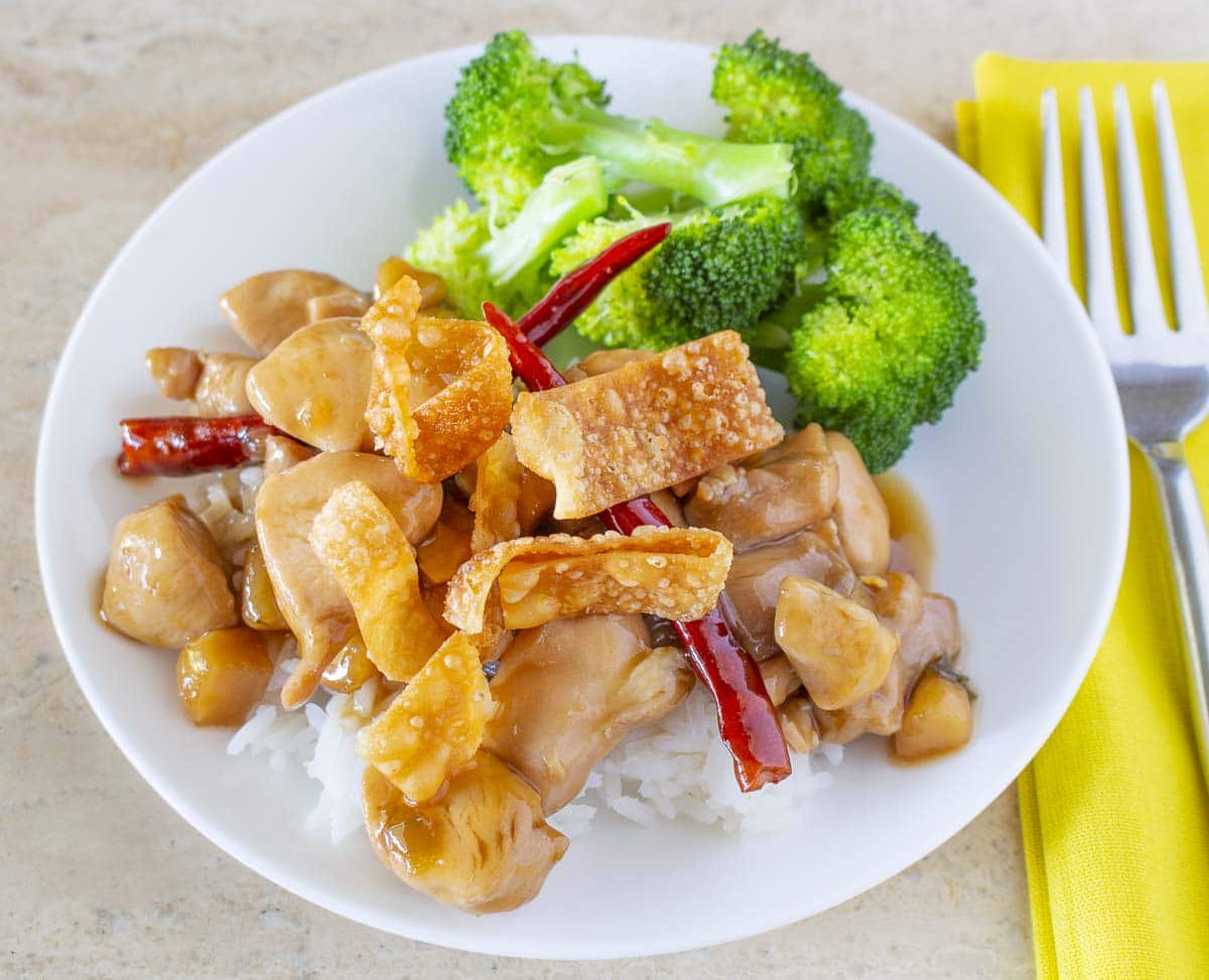 Chicken in spicy sauce with dried red peppers topped with fried wonton strips over rice on a plate with streamed broccoli