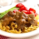 Beef tips in gravy over noodles on a plate with tomatoes and asparagus