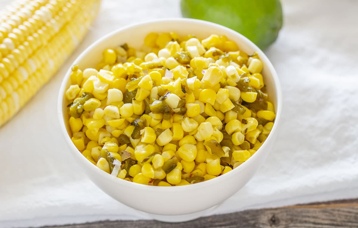 Corn salsa in a white bowl with fresh corn and lime in the background