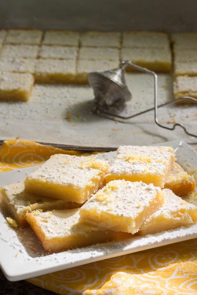 Lemon bars with powdered sugar and lemon zest in front of cut bars and a flour duster