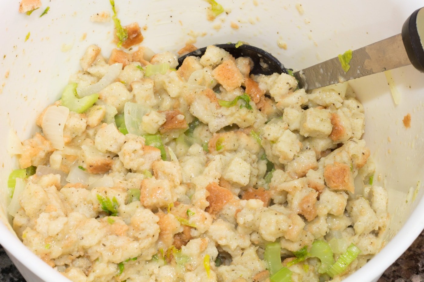 Moistened bread cubes with celery and onions in a white bowl with a mixing spoon