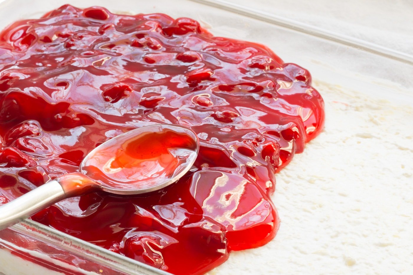 Cherry pie filling being spooned onto cherry delight pineapple cream cheese base in a white baking dish