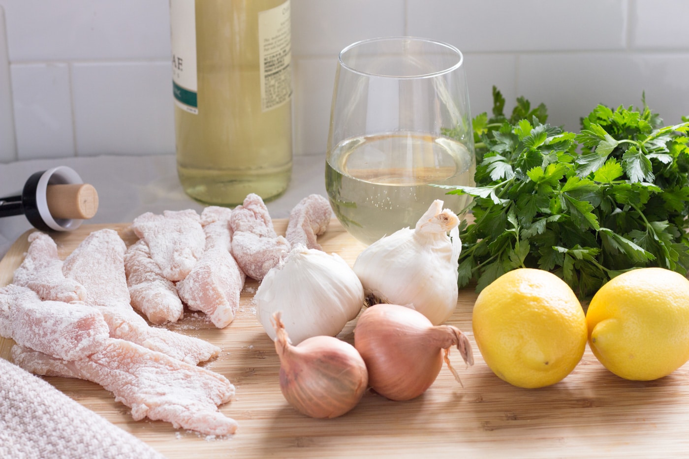 floured chicken strips shallots garlic lemons parsley and a glass of wine on a cutting board