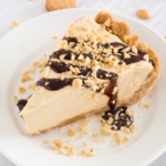 Slice of peanut butter pie with crushed peanuts and chocolate syrup on a white plate with mini nutter butters around it