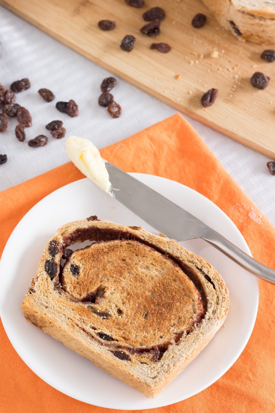 Toasted cinnamon raisin bread and butter knife on a white plate sitting on an orange napkin