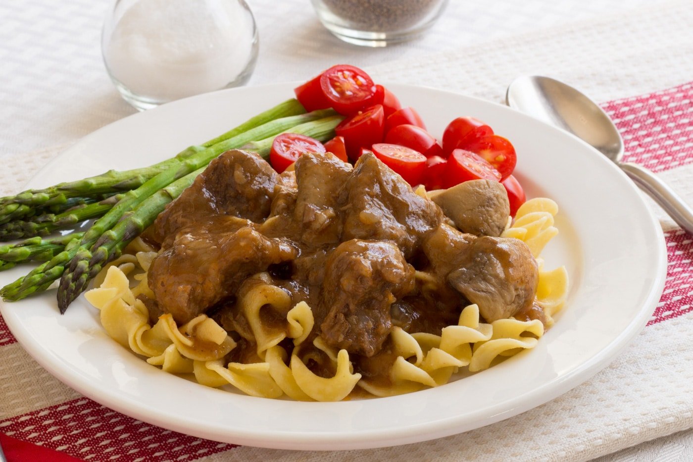 beef tips with gravy over noodles on white plate with tomatoes and asparagus