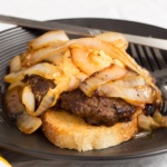 Sour Daough Parmesan toast topped with a fried burger pub cheese abd grilled onions on a black plate with a fork and knife