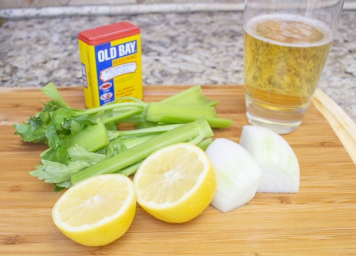 Lemon halves, onions wedges, celery pieces, Old Bay seasoning, and a glass of beer on a cutting board