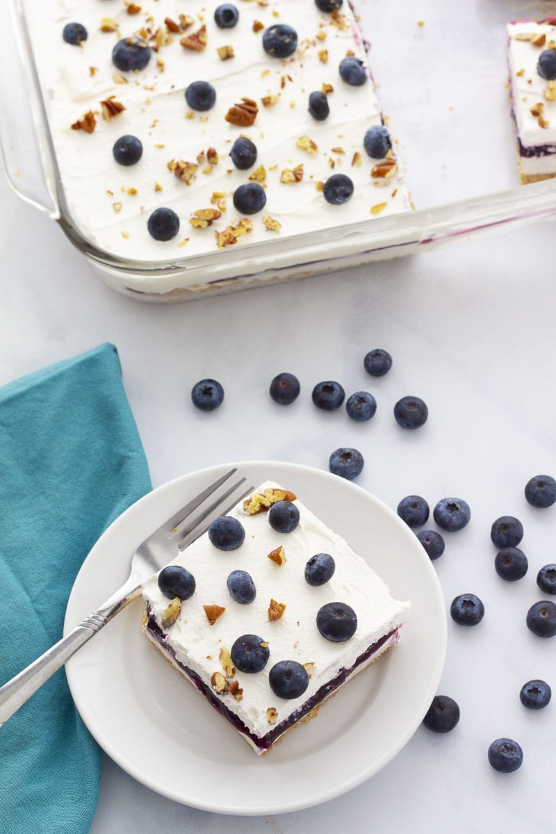 Slice of dessert covered with fresh blueberries and nuts on a plate and a full pan in the back.