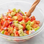 Diced cucumbers and tomatoes covered in dressing in a bowl with a wooden salad spoon
