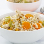 White bowl with couscous, chicken, celery, and carrots with an orange check napkin and spoons in the background