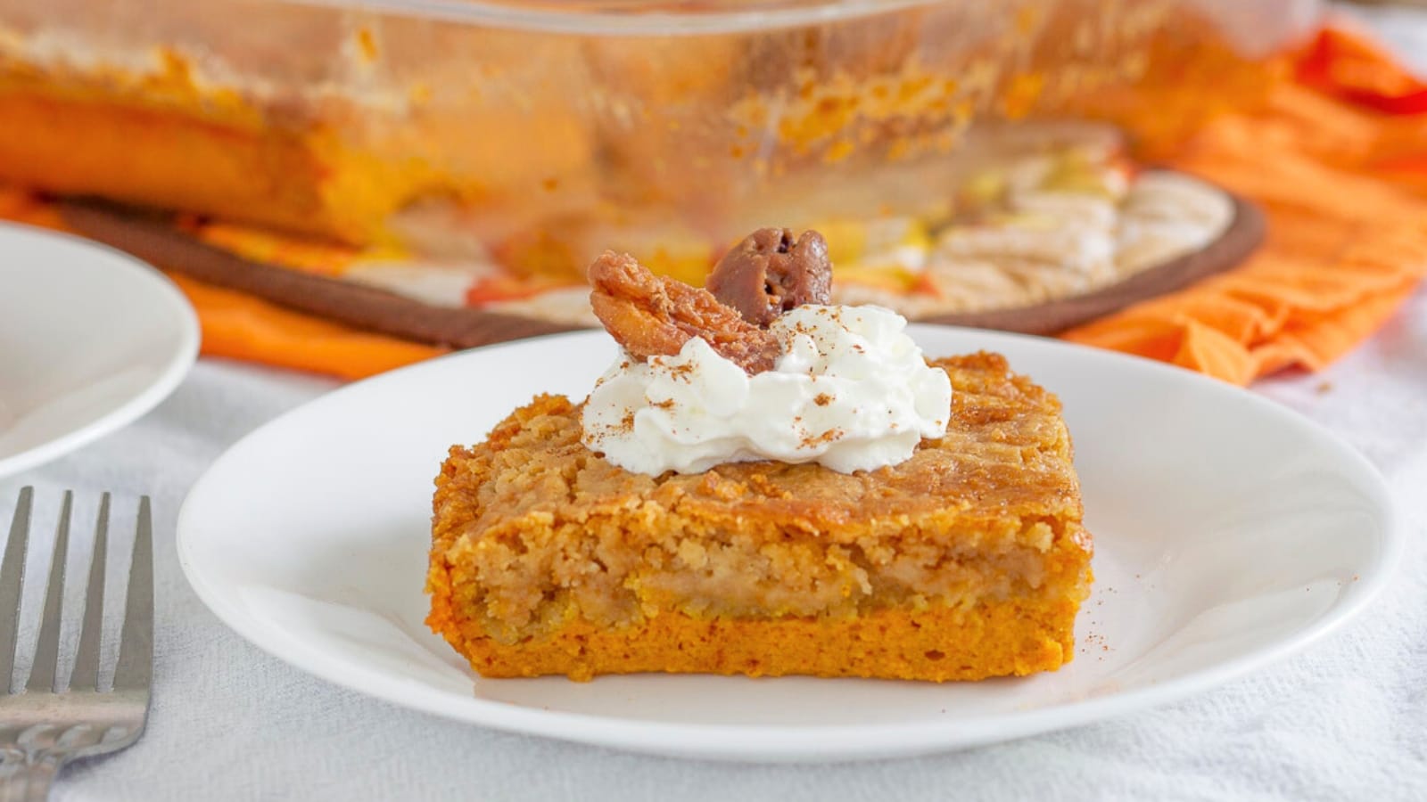 Slice of pumpkin dump cake topped with whipped cream, cinnamon sprinkles, and candied pecans on a plate next to a fork in from of the glass baking dish