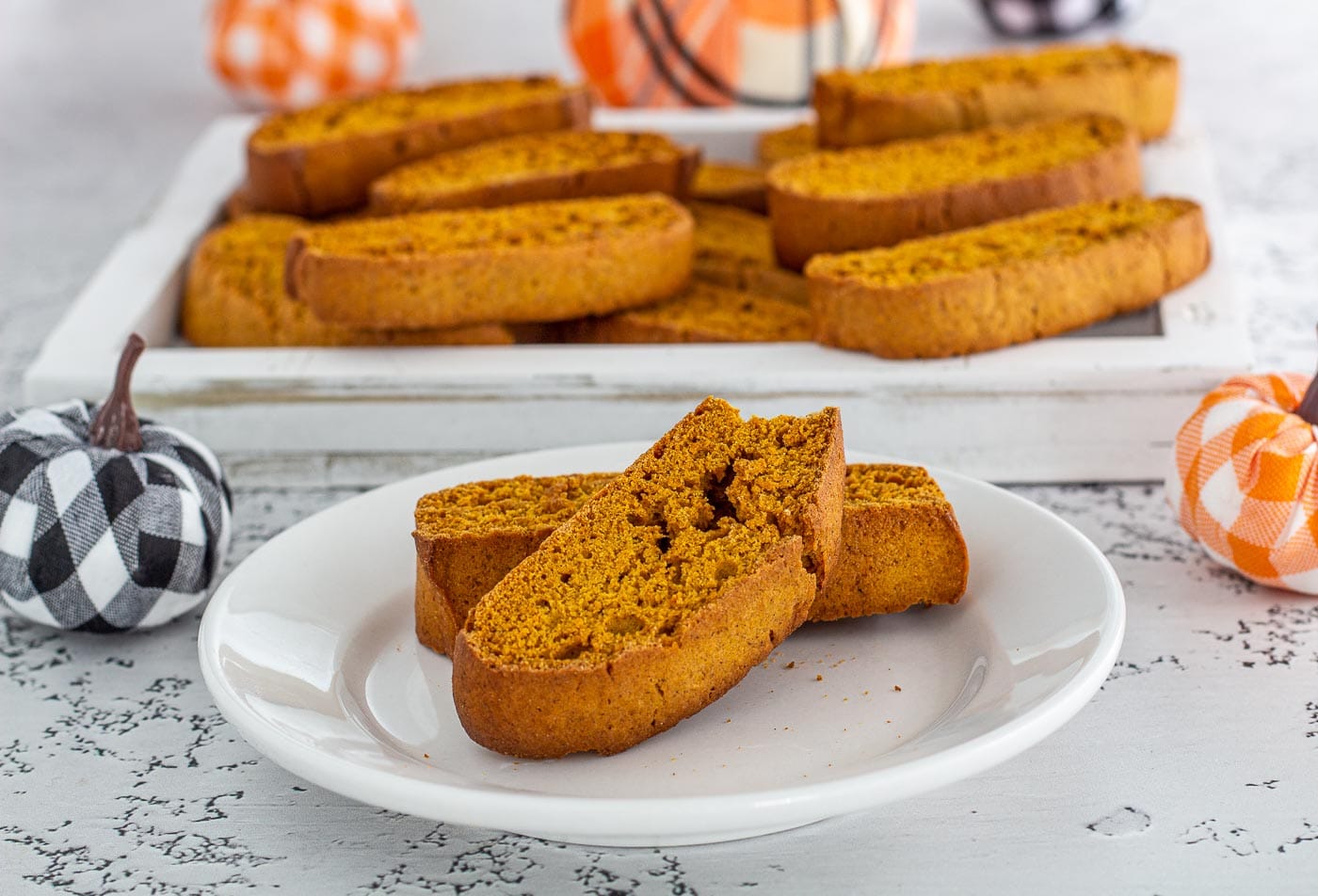 Pumpkin biscotti on a plate in front of a tray of cookies and 2 plaid pumpkins