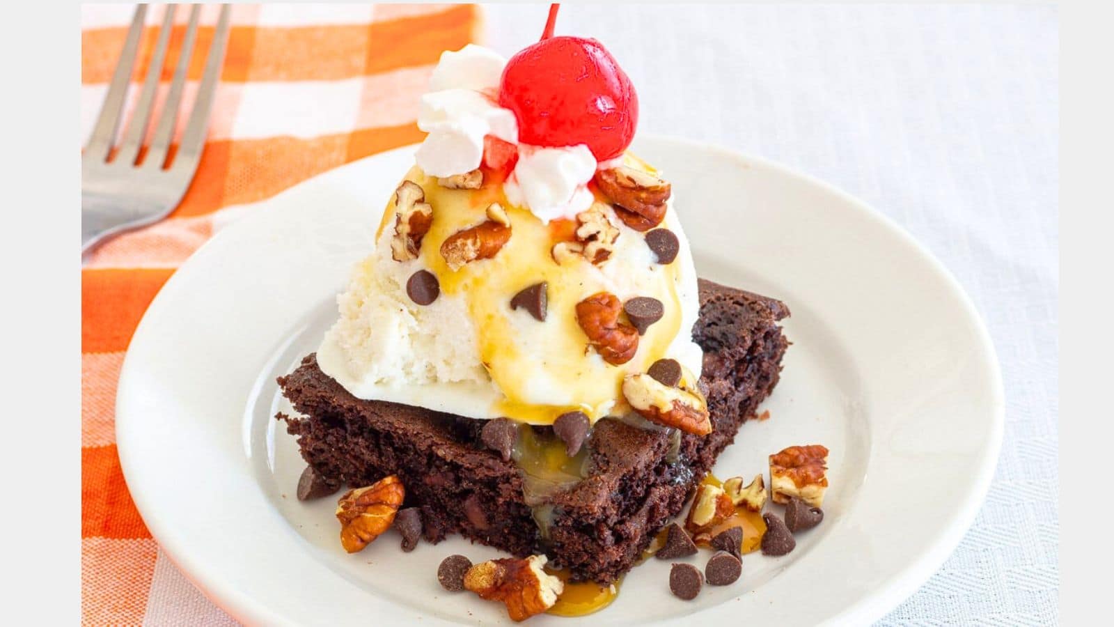 Chocolate cake topped with ice cream chocolate chips nuts and a cherry on a plate with an orange checked napkin