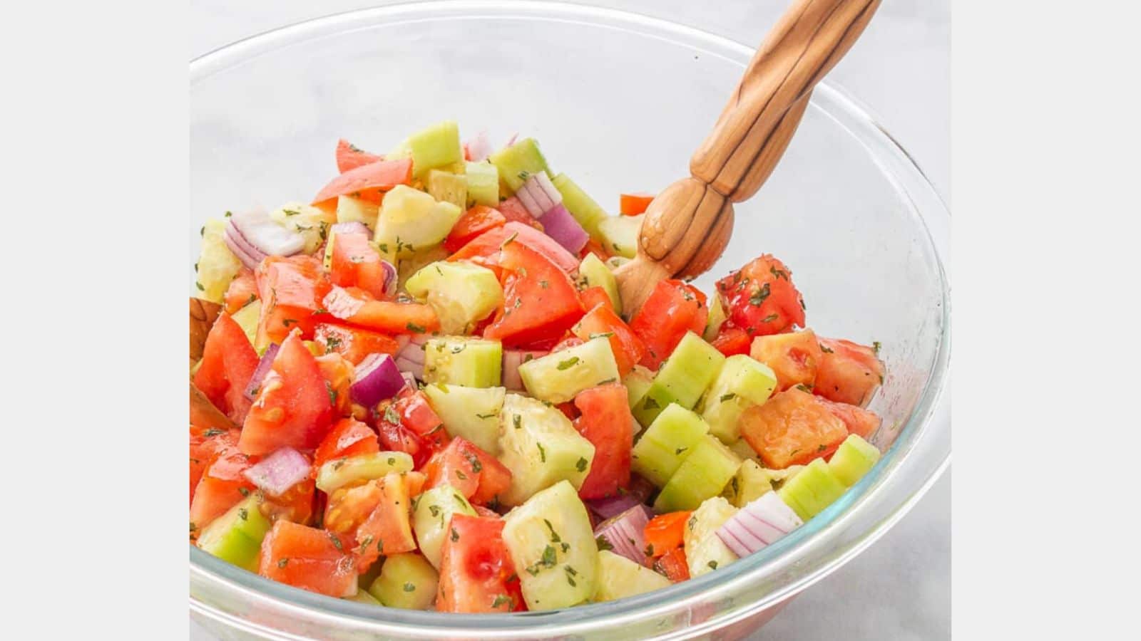 Cucumber and tomato salad in a glass bowl with a wooden spoon