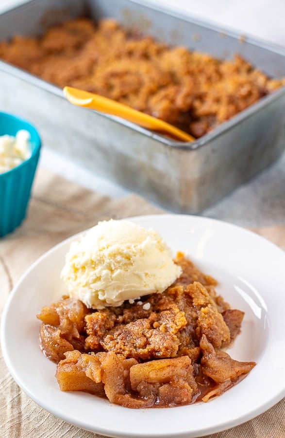 Slice of apple crumble topped with ice cream on a plate with a blue bowl of ice cream and a pan of apple crumble in the background