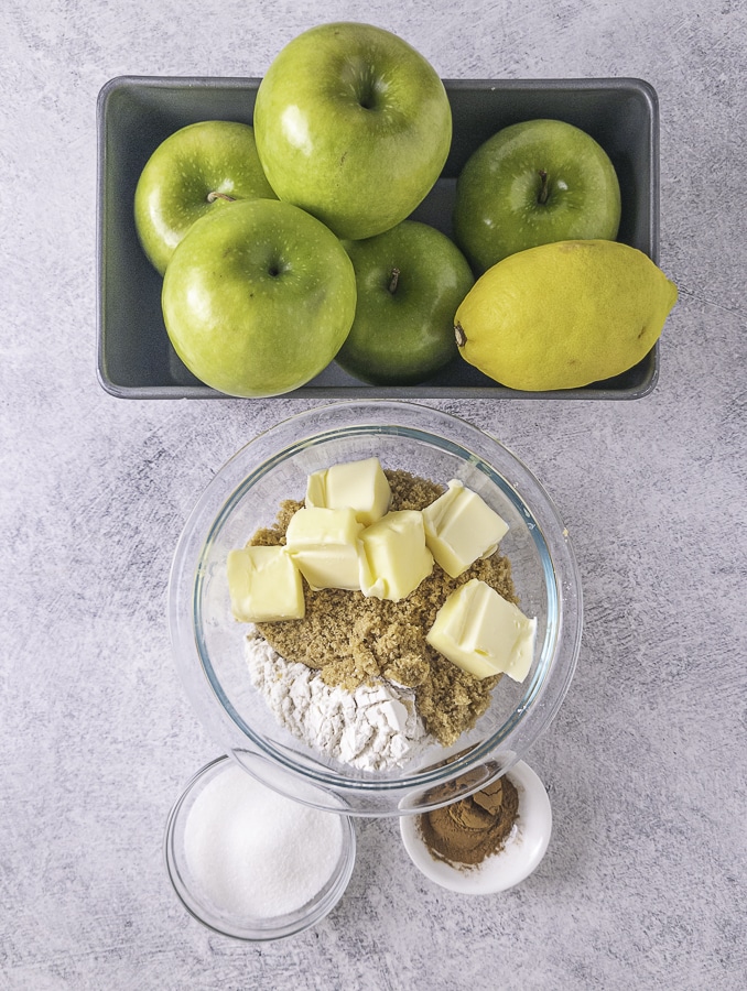 Green apples and a lemon in a pan a bowl with flour butter and brown sugar with white sugar and cinnamon in bowls at the bottom