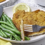chicken cutlet with a bite on a fork green beans a lemon slice and knife on a palte
