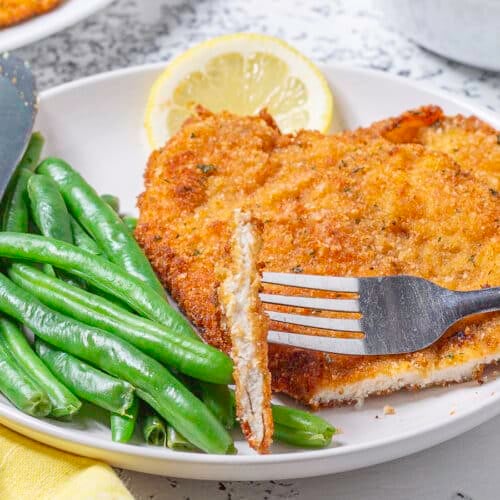 chicken cutlet with a bite on a fork green beans a lemon slice and knife on a palte