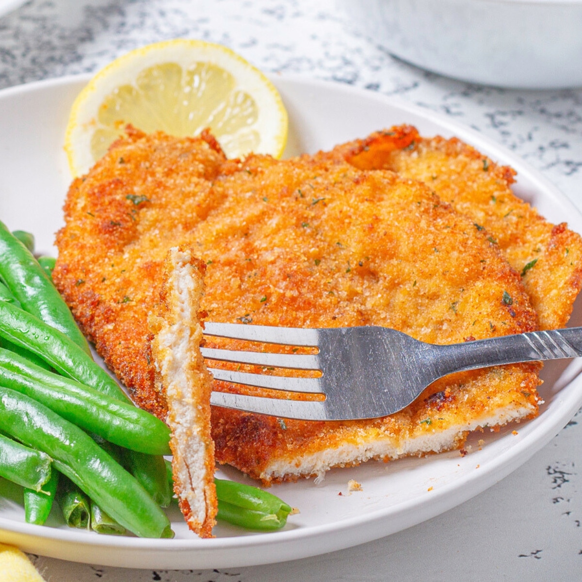 pan fried chicken cutlet with a bite on a fork sitting on a plate with green beans and a lemon slive