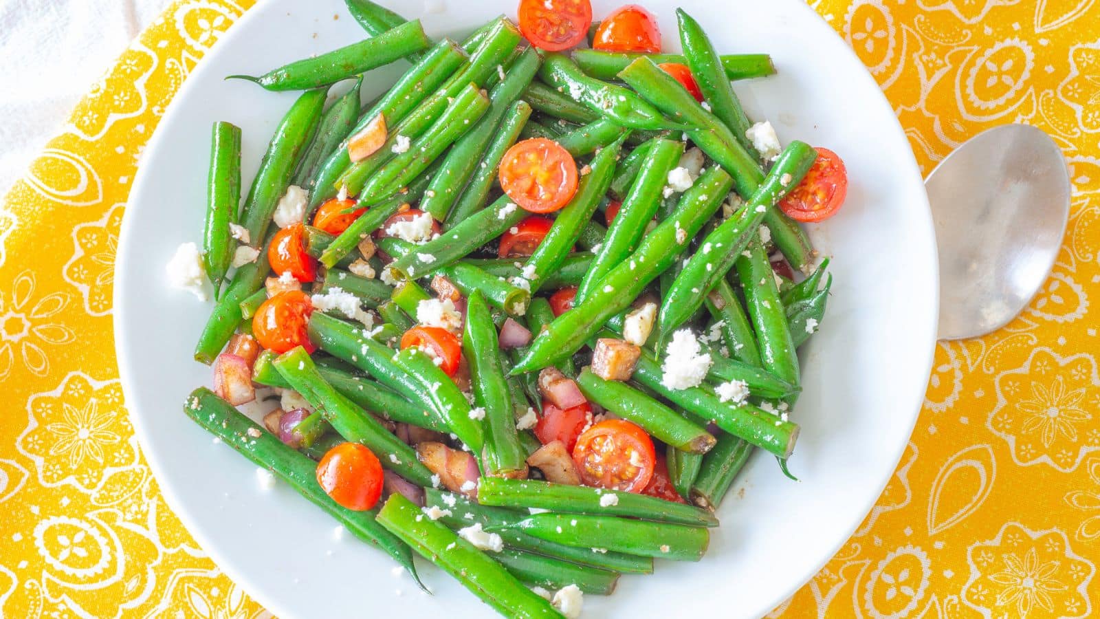 Green beans with tomatoes and cheese crumbles in a bowl next to a spoon