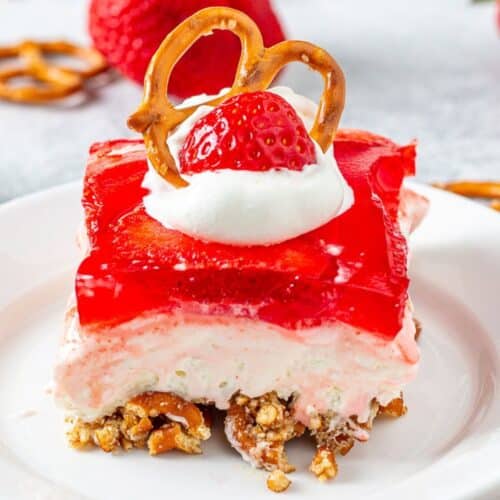 Slice of strawberry pretzel salad on a plate topped with a pretzel and strawberry piece on a plate surrounded by pretzels and strawberries