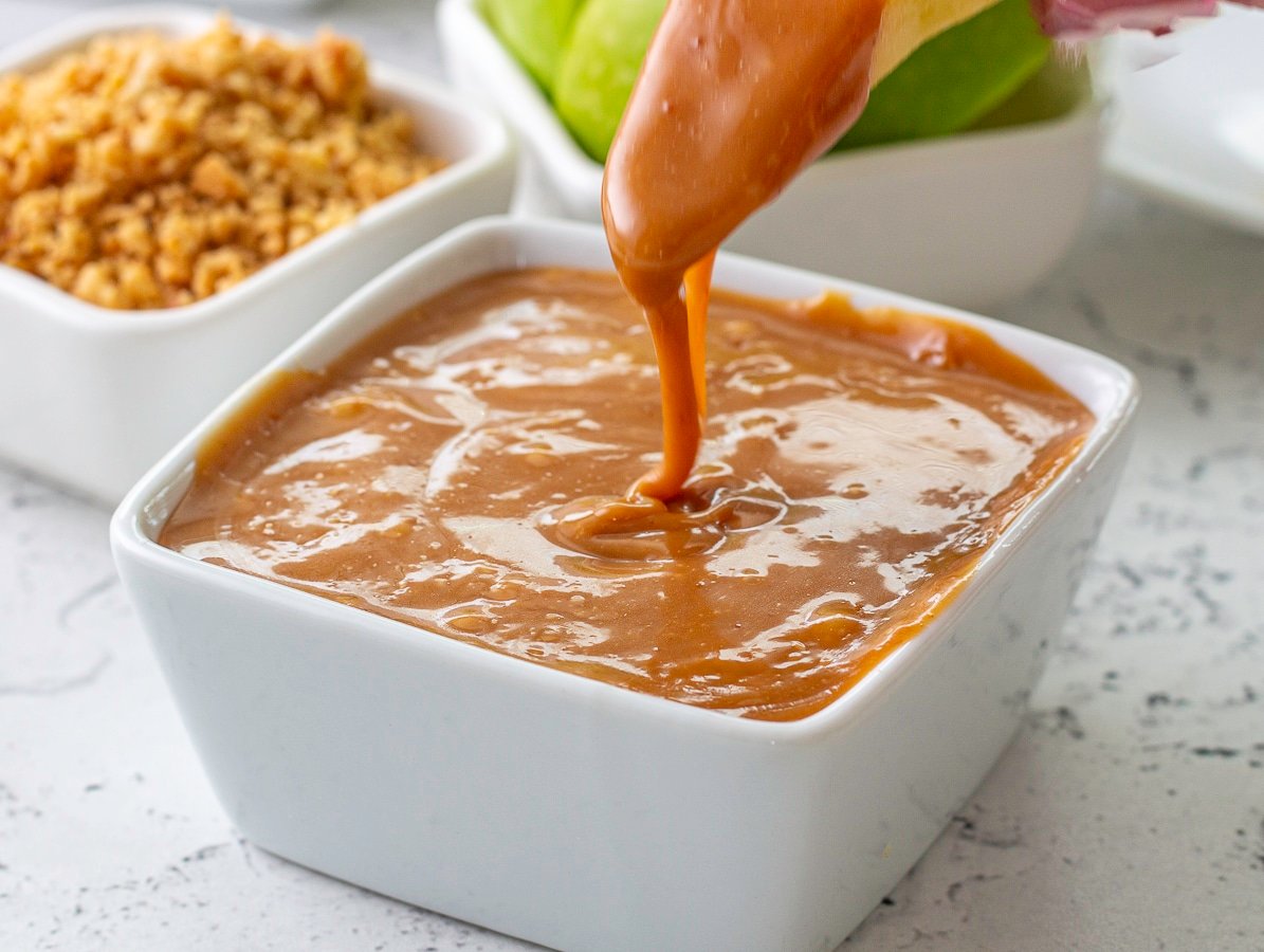 Dipping an apple slice into caramel sauce with peanuts and apples in the background