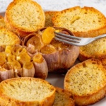 Roasted garlic clove on a fork above 2 heads of roasted garlic and crostini