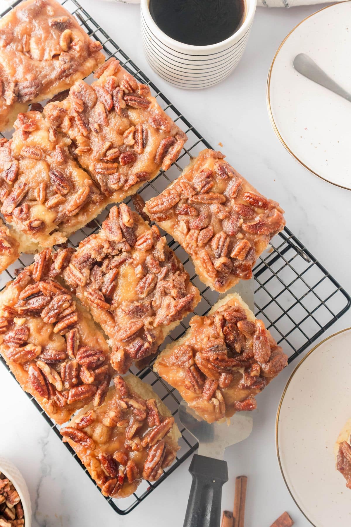 Breakfast rolls topped with caaramel and pecans on a cooling rack next to a cup of coffee