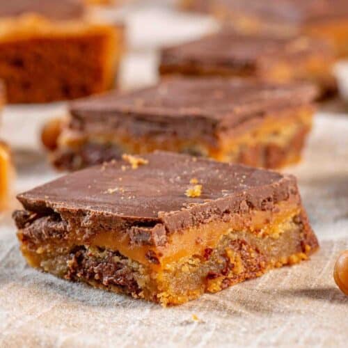 Chocolate chip cookie bars topped with caramel and chocolate lined up on a beige napkin