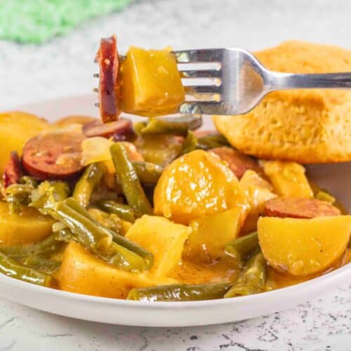 potatoes green beans and sausage on a plate with a biscuit with a fork holding a piece of sausage and potato chunk