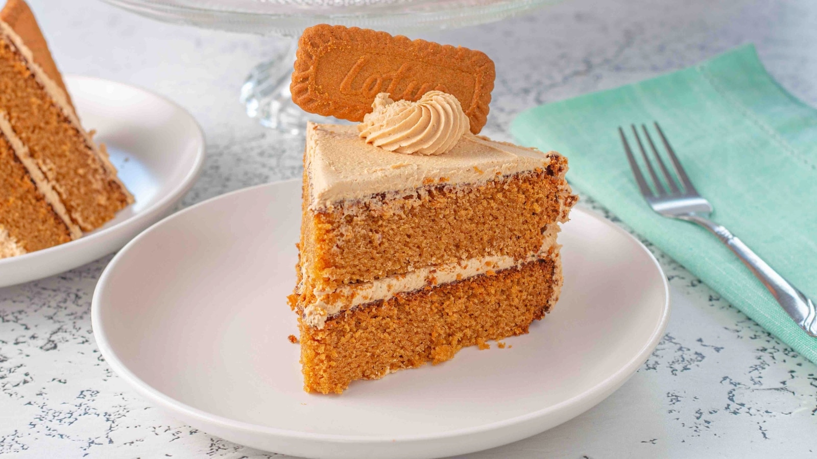 Slice of cookie butter cake topped with a frosting swirl and a full cookie on a white plate sitting next to a teal napkin and fork